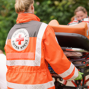 DRK-Einsatzkraft in orangefarbener Schutzjacke mit weißem Kreuz auf dem Rücken schiebt eine Rettungstrage durch eine grüne Umgebung.
